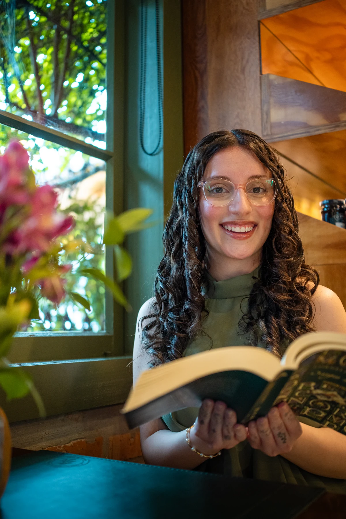 Giulia Campos sorrindo, com um livro aberto nas mãos, ao lado de uma janela